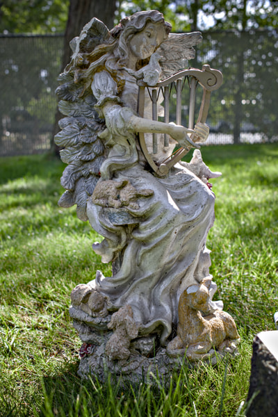 A weathered stone angel statue holding a lyre watches over lamb figures at Pilot Butte Cemetery.